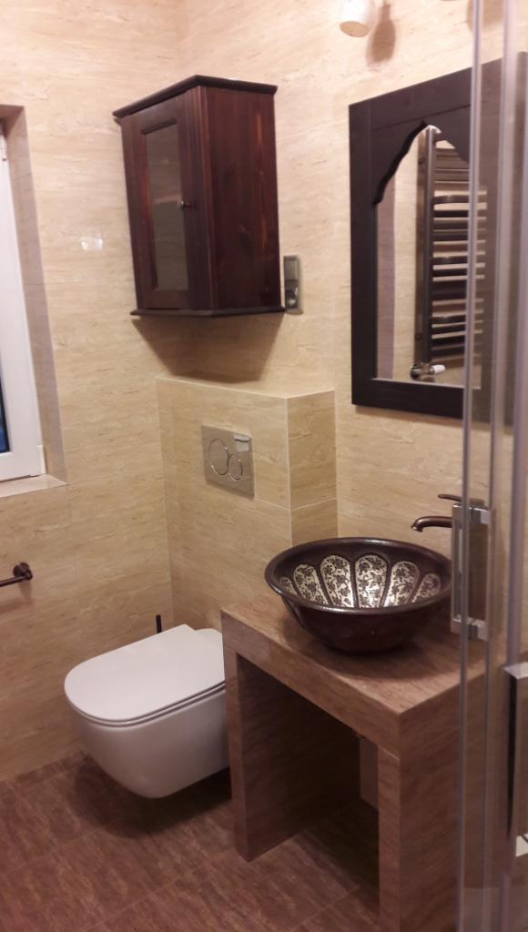 Nowoczesna łazienka z podwieszaną toaletą, umywalką w stylu orientalnym i drewnianą szafką wiszącą, wykończona beżowymi płytkami imitującymi trawertyn.