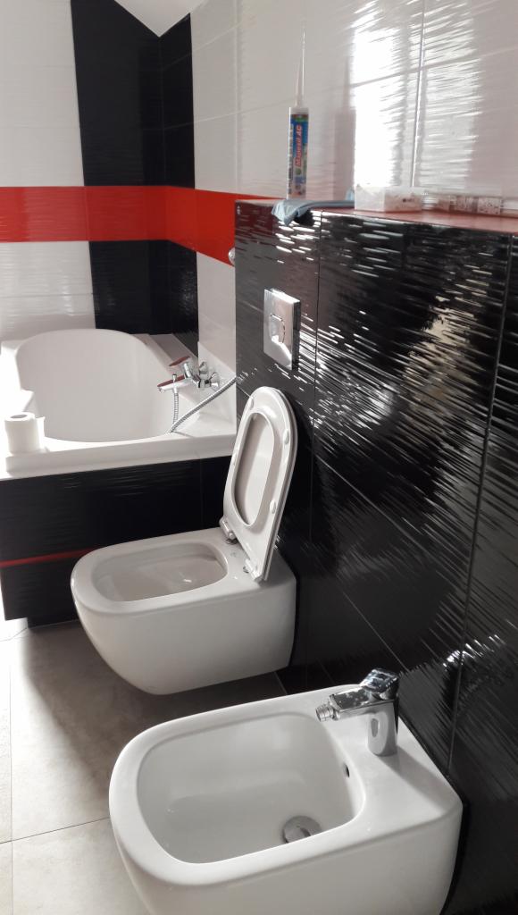 Nowoczesna łazienka z białą armaturą: toaleta z otwartą klapą i bidetem, na tle czarno-biało-czerwonych płytek oraz akcesoria montażowe na blacie.