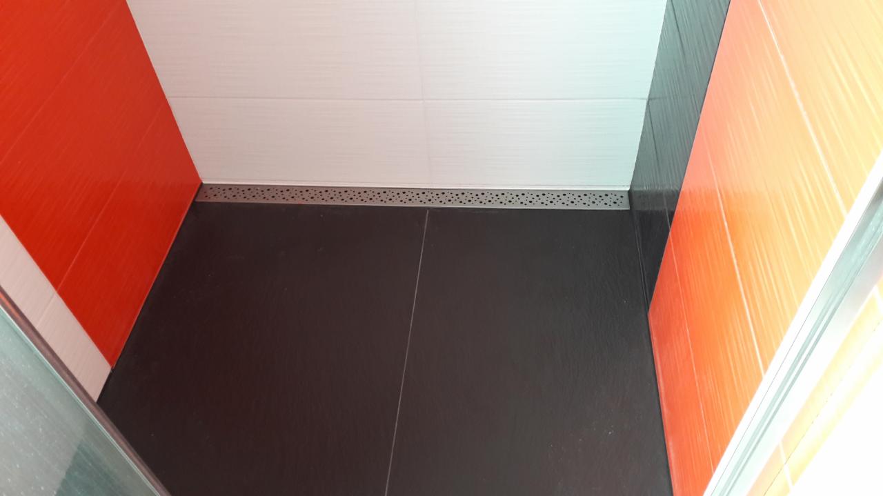 Nowoczesna kabina prysznicowa z czarną podłogą, białymi i pomarańczowymi płytkami ściennymi oraz odpływem liniowym z perforowanym rusztem.