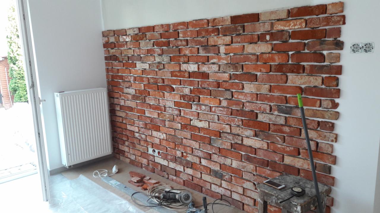 Ściana wyłożona dekoracyjną cegłą w odcieniach czerwieni i brązu, widoczne narzędzia pracy, szlifierka kątowa i poziomica, fragment grzejnika i otwarte drzwi balkonowe.
