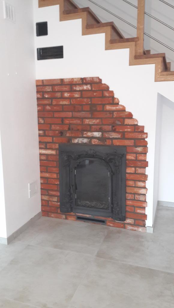 Częściowo wykończona ściana z czerwonej cegły wokół czarnego, ozdobnego wkładu kominkowego, z widocznymi schodami z drewnianymi stopniami w tle.