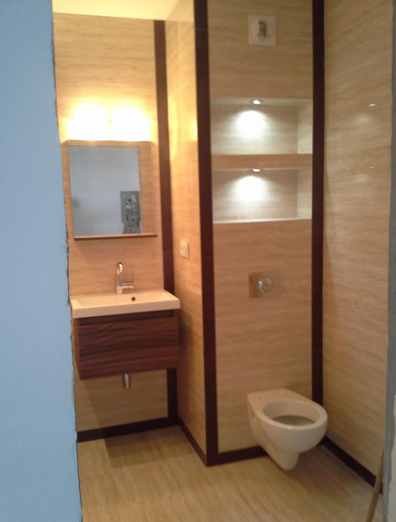 Wykończona łazienka z beżowymi płytkami imitującymi drewno, wisząca szafka z umywalką, toaleta podtynkowa i wnęka z półkami oświetlona punktowymi lampkami.