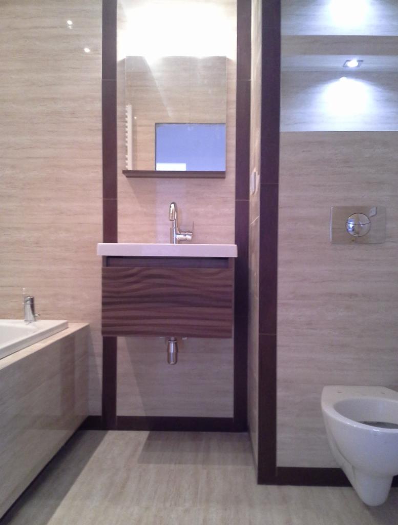 Nowoczesna łazienka z beżowymi płytkami imitującymi drewno, wisząca szafka z umywalką o froncie z ciemnego drewna z wyraźnym usłojeniem, lustro z minimalistyczną półką, podwieszana toaleta...