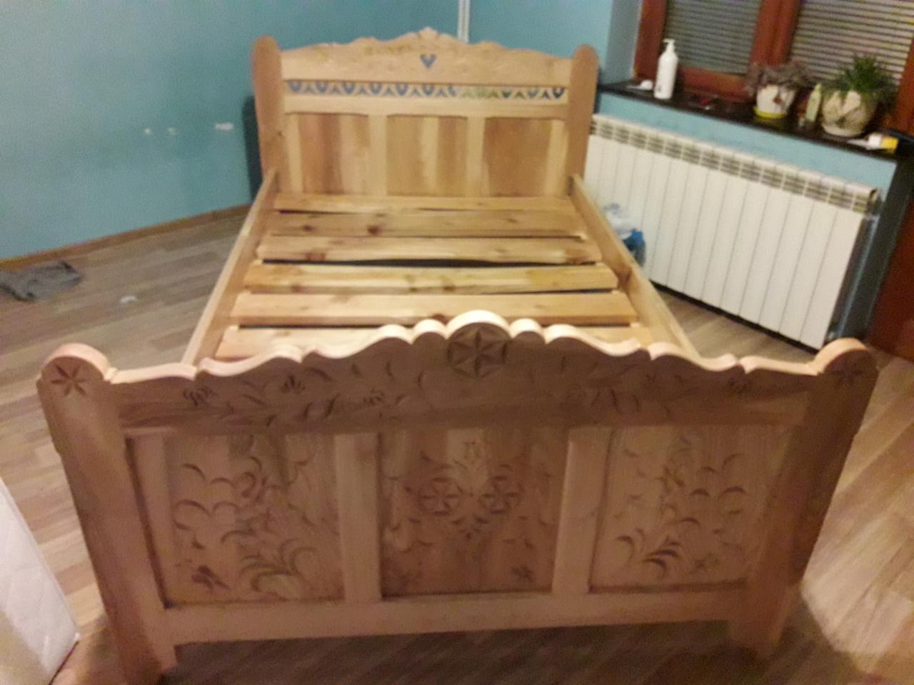 Surowe drewniane łóżko z dekoracyjnymi rzeźbieniami na wezgłowiu i zanóżku, widoczne listwy pod materac, w tle ściana w kolorze turkusowym i grzejnik.