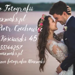 Para młoda, panna młoda w koronie z kwiatów i sukni z koronkowymi rękawami, pan młody w granatowym garniturze, obejmująca się na tle rozmytego krajobrazu, logo studia fotograficznego w lewym górnym...