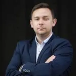 Portret mężczyzny w niebieskiej marynarce z założonymi rękami na tle ciemnej ściany. Widoczny zegarek na lewym nadgarstku.