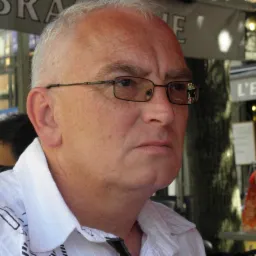 Portret mężczyzny w okularach, zamyślony wyraz twarzy, w tle kawiarnia L'Esquille.