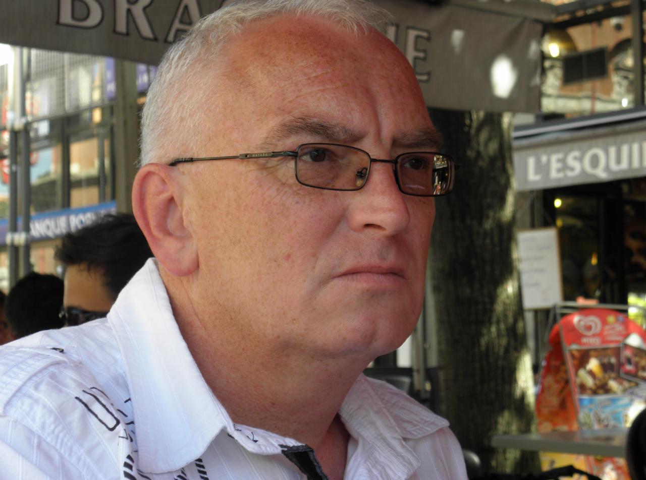 Portret mężczyzny w okularach, zamyślony wyraz twarzy, w tle kawiarnia L'Esquille.