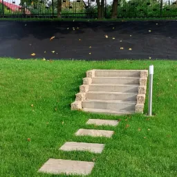 Betonowe schody z kamiennymi obrzeżami prowadzące przez trawnik, z pojedynczymi kamiennymi płytami tworzącymi ścieżkę, zakończone brukowaną powierzchnią, na tle drzew i czarnego muru oporowego.
