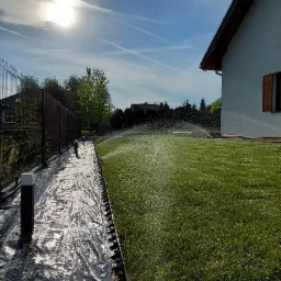 Automatyczny system nawadniania trawnika w trakcie pracy, widok z poziomu gruntu na zraszacze, trawnik i obrzeże z folią ochronną.