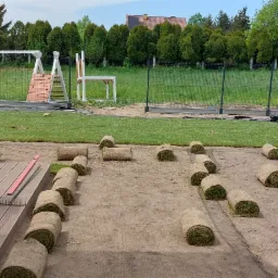 Rozwinięte rolki trawy na przygotowanym podłożu, widoczny pracownik oraz narzędzia na drewnianym tarasie, w tle dom i plac zabaw.