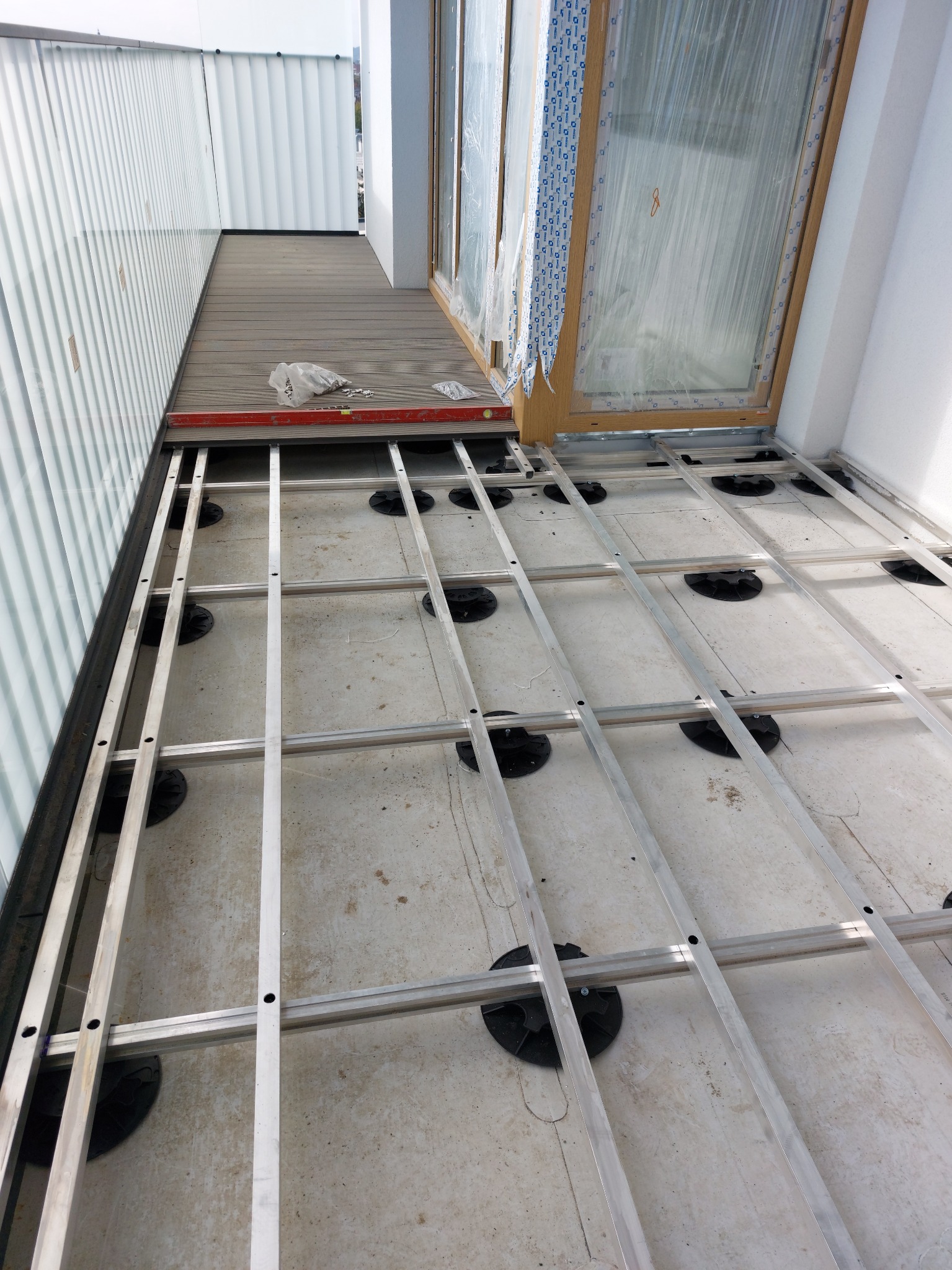 Montaż konstrukcji pod taras kompozytowy: aluminiowe legary na regulowanych wspornikach, częściowo ułożona deska tarasowa, poziomica i akcesoria na górze.