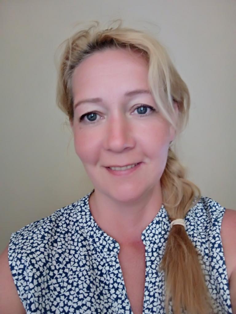 Portret uśmiechniętej kobiety z blond włosami spiętymi w warkocz, ubrana w bluzkę w biało-granatowe wzory na jednolitym, jasnym tle.