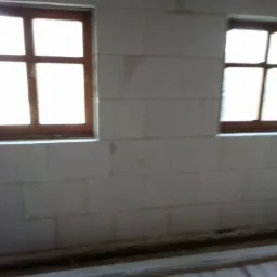 Ściana z białych bloczków z dwoma oknami z brązowymi ramami, widoczne ślady po zaprawie, surowy stan wykończenia.