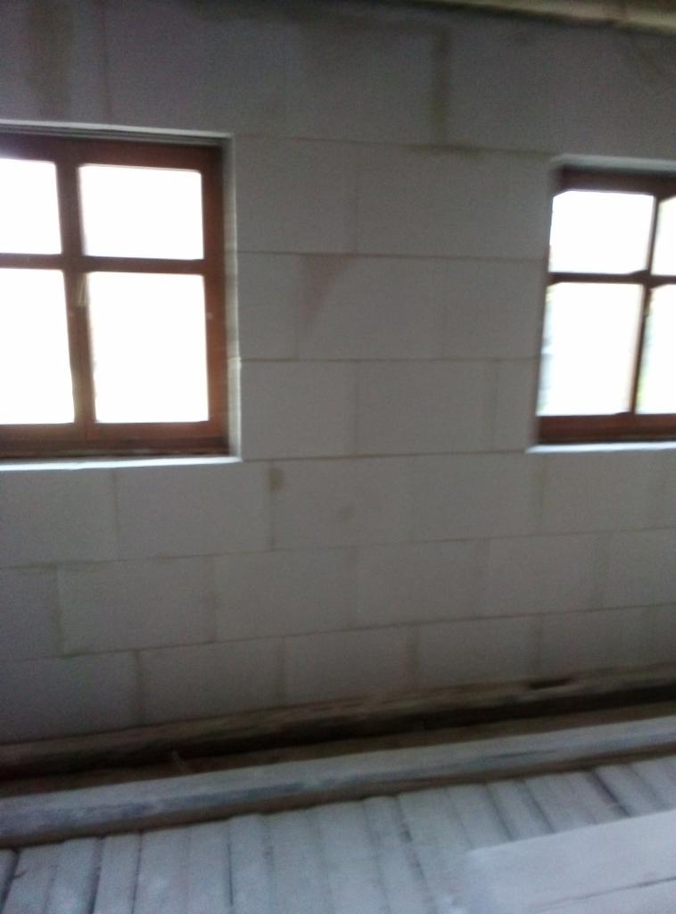 Ściana z białych bloczków z dwoma oknami z brązowymi ramami, widoczne ślady po zaprawie, surowy stan wykończenia.