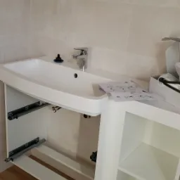 Montaż umywalki z szafką w łazience, widoczne elementy mocujące szuflady, instrukcja na blacie i syfon w szafce.