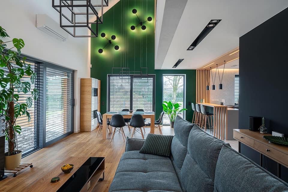 Nowoczesny salon z zieloną ścianą, drewnianą podłogą i szarą sofą; widoczne geometryczne lampy sufitowe, stół z krzesłami oraz roślina doniczkowa