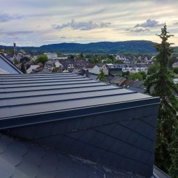 Dekarz Cieśla Usługi Ogólnobudowlane - Widok na miasto z nowo położonego, ciemnego dachu z blachy trapezowej i łupka, w tle zielone wzgórza i pochmurne niebo.
