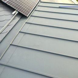 Dekarz Cieśla Usługi Ogólnobudowlane - Szary dach z blachy na rąbek stojący, z zamontowanymi panelami słonecznymi i szczotką do mycia na długim kiju.