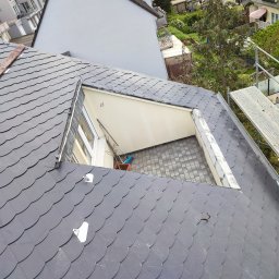 Dekarz Cieśla Usługi Ogólnobudowlane - Fragment dachu pokrytego łupkiem z otwartym oknem dachowym, widoczne rusztowanie i elementy mocujące łatę dachową.
