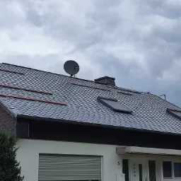 Dom jednorodzinny z dachem pokrytym ciemnoszarą łupkową dachówką, widoczne okna dachowe, antena satelitarna i fragment elewacji z numerem domu.