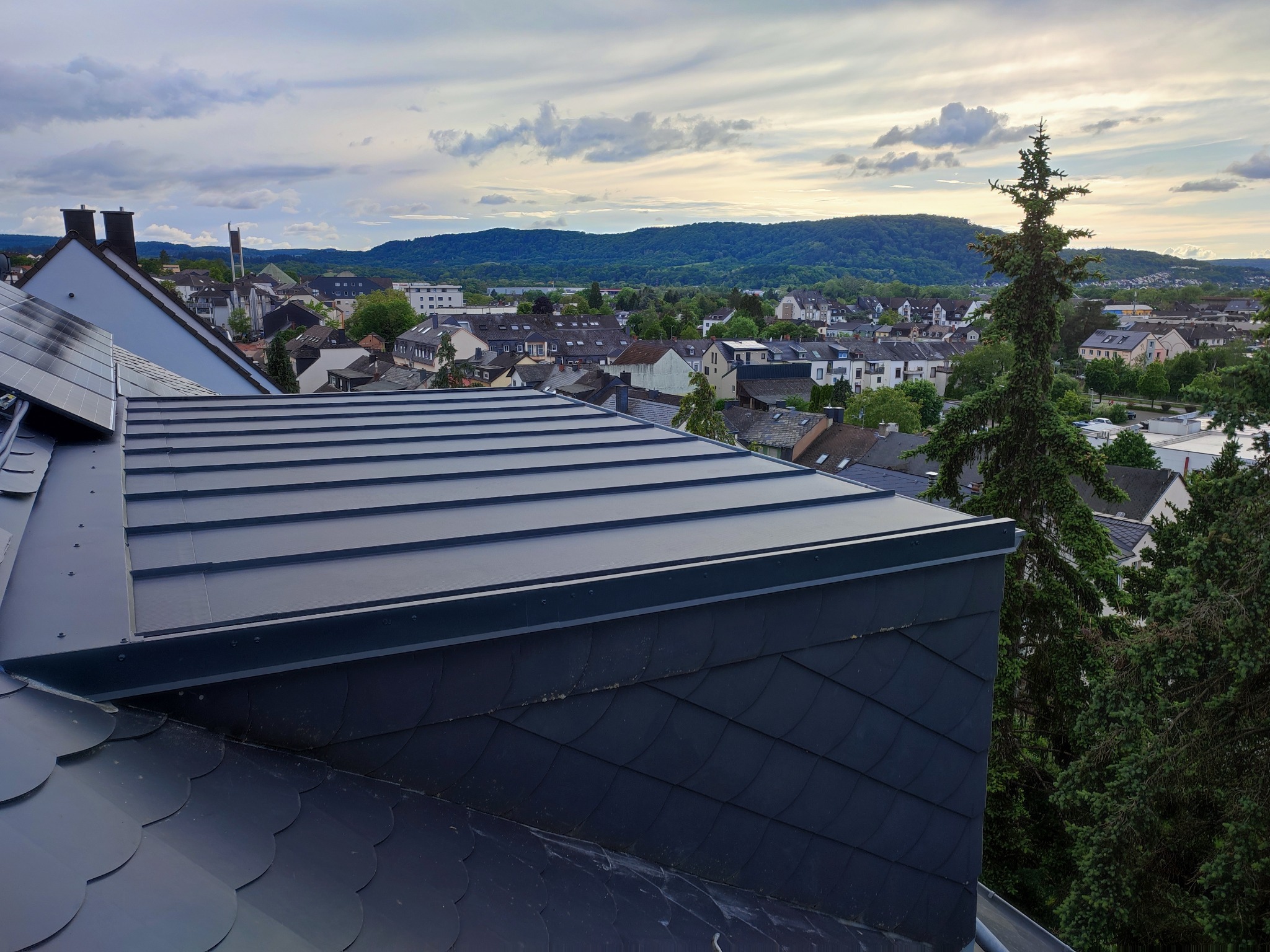 Widok na miasto z nowo położonego, ciemnego dachu z blachy trapezowej i łupka, w tle zielone wzgórza i pochmurne niebo.