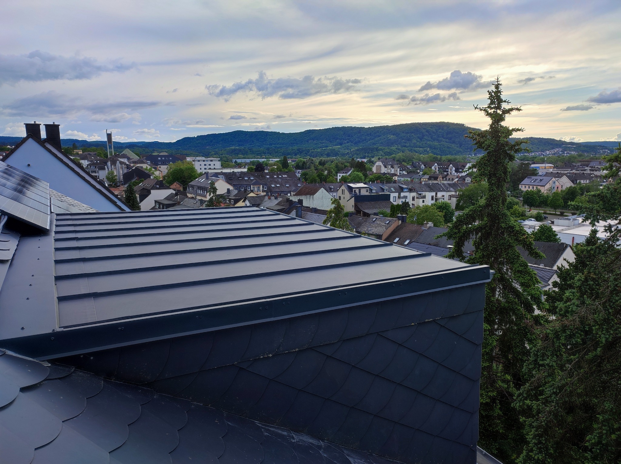 Nowoczesne krycie dachu z blachy na rąbek stojący w kolorze grafitowym, widok na panoramę miasta i zalesione wzgórza w tle.