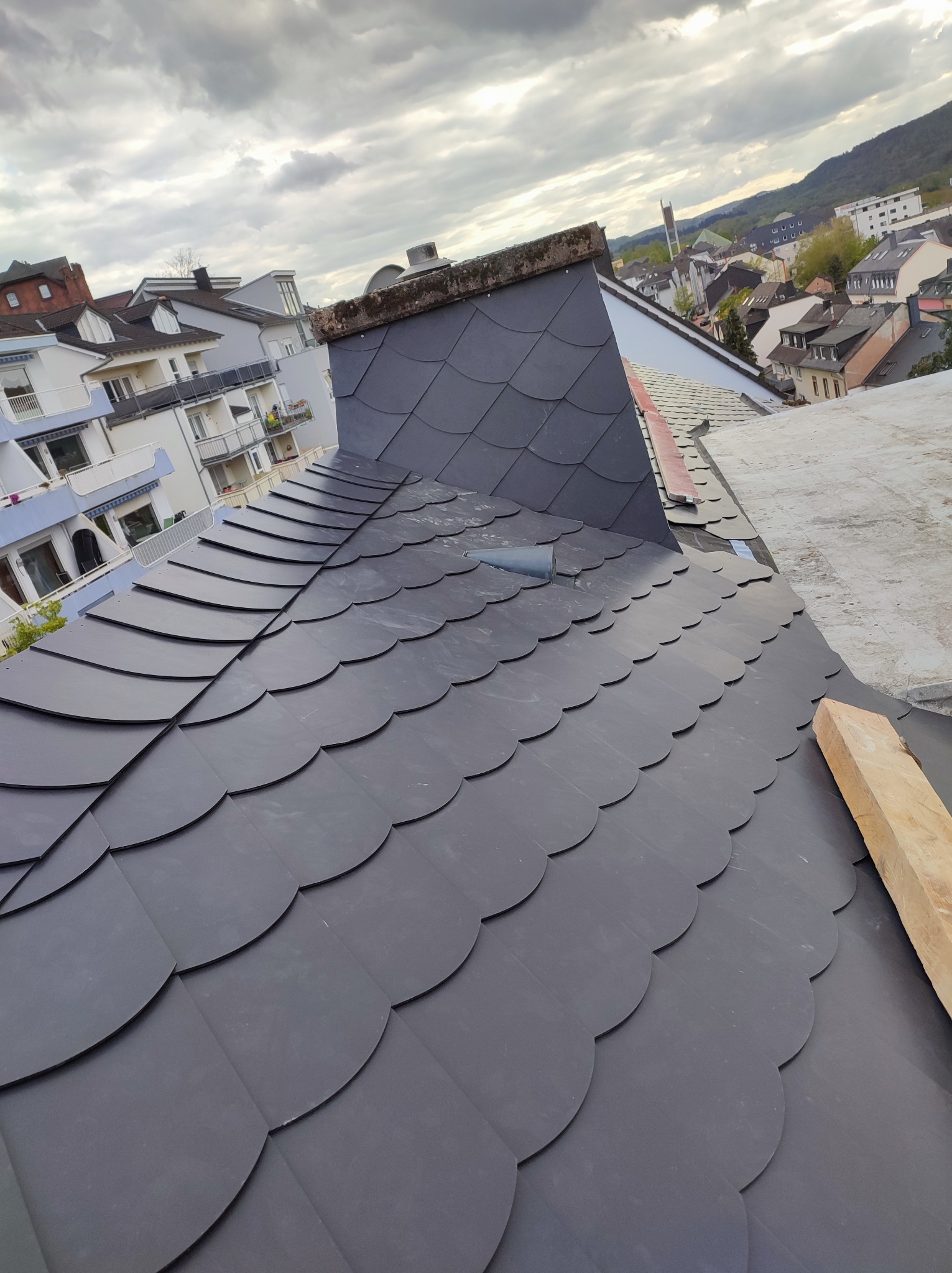 Nowy, ciemnoszary dach pokryty łuską bitumiczną, widok z góry na tle zabudowy miejskiej i pochmurnego nieba.
