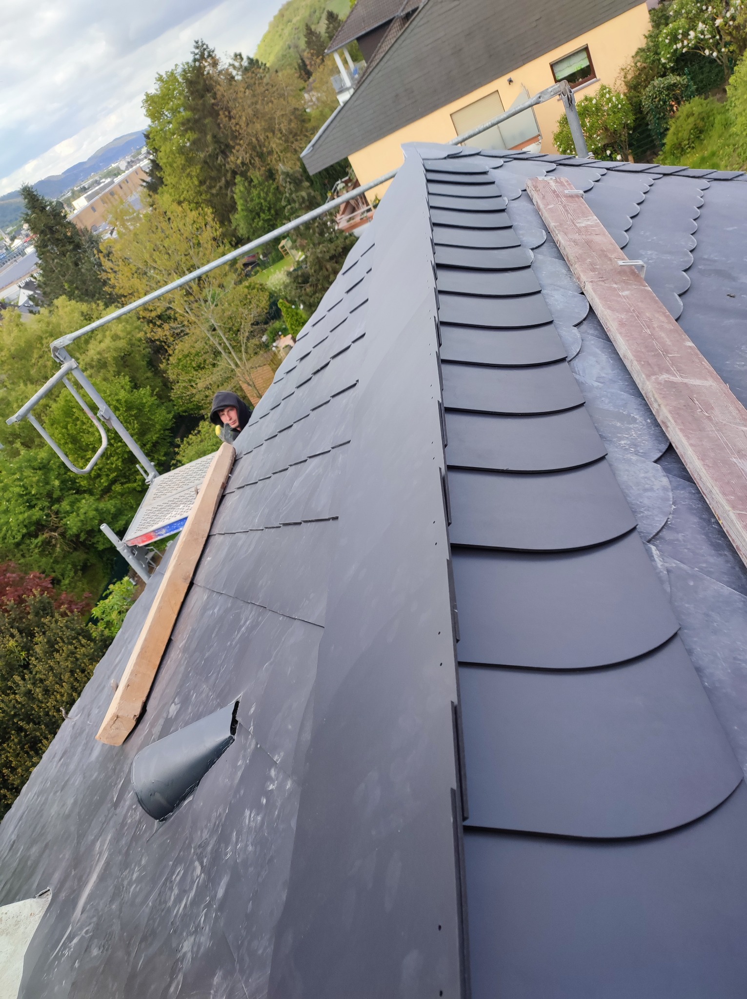 Układanie ciemnoszarej dachówki karpiówki na dachu z widocznym fragmentem rusztowania i pracownikiem w tle, deski pomocnicze na powierzchni dachu.