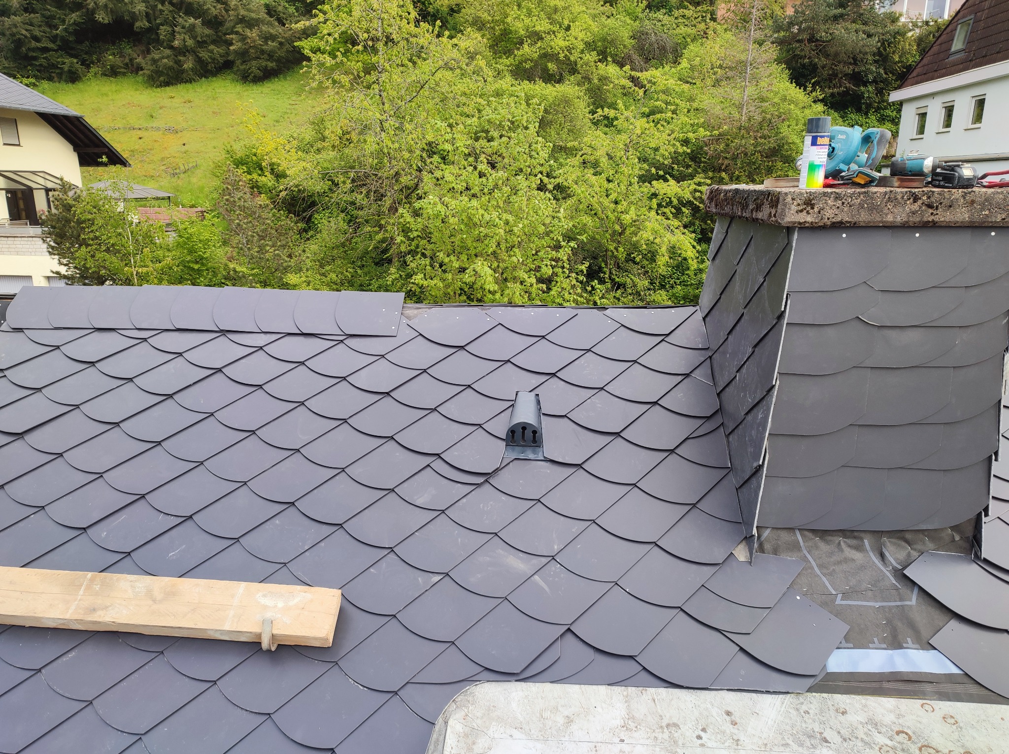 Nowy dach z łupka w trakcie układania, widoczny komin, narzędzia i fragment zielonego wzgórza w tle.