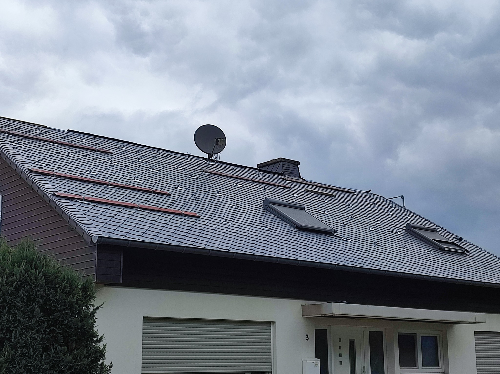 Dom jednorodzinny z dachem pokrytym ciemnoszarą łupkową dachówką, widoczne okna dachowe, antena satelitarna i fragment elewacji z numerem domu.