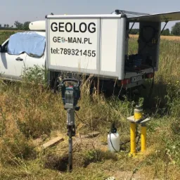Prace terenowe: Wiercenie otworu w ziemi w otoczeniu wysokich traw, widoczny sprzęt do wiercenia i samochód dostawczy z napisem GEOLOG