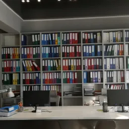 Biuro z regałem pełnym kolorowych segregatorów, biurko z komputerami i lampami, drukarka w rogu pokoju.