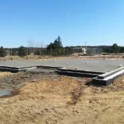 Wylany, betonowy fundament domu jednorodzinnego z hydroizolacją, widok na teren budowy z piaskiem i śladami opon, w tle zalesiony krajobraz pod błękitnym niebem.