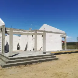 Surowy stan domu murowanego z betonu kom&oacute;rkowego Ytong, widoczne ściany, betonowe słupy i schody wejściowe, w tle błękitne niebo.