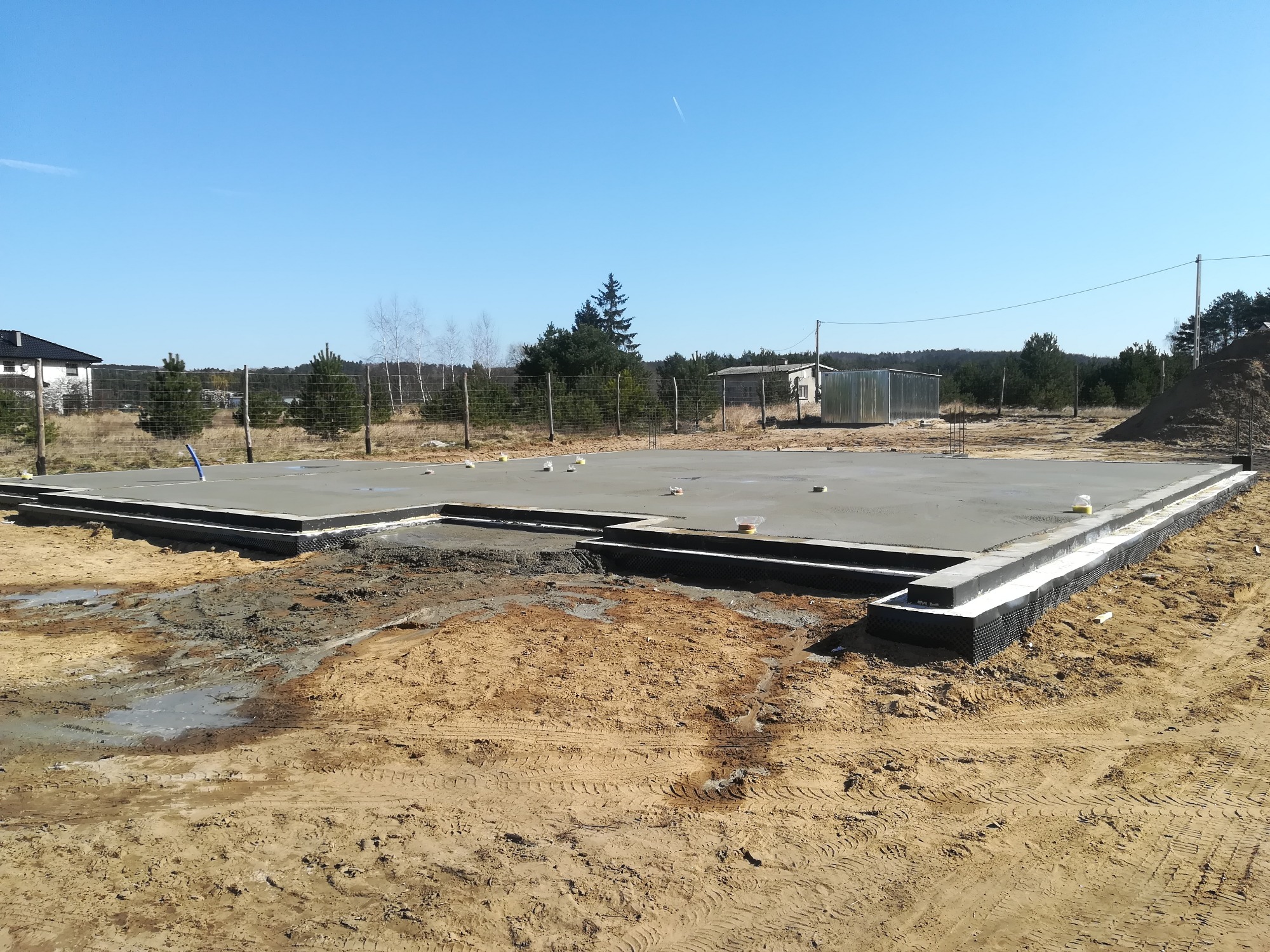 Wylany, betonowy fundament domu jednorodzinnego z hydroizolacją, widok na teren budowy z piaskiem i śladami opon, w tle zalesiony krajobraz pod błękitnym niebem.
