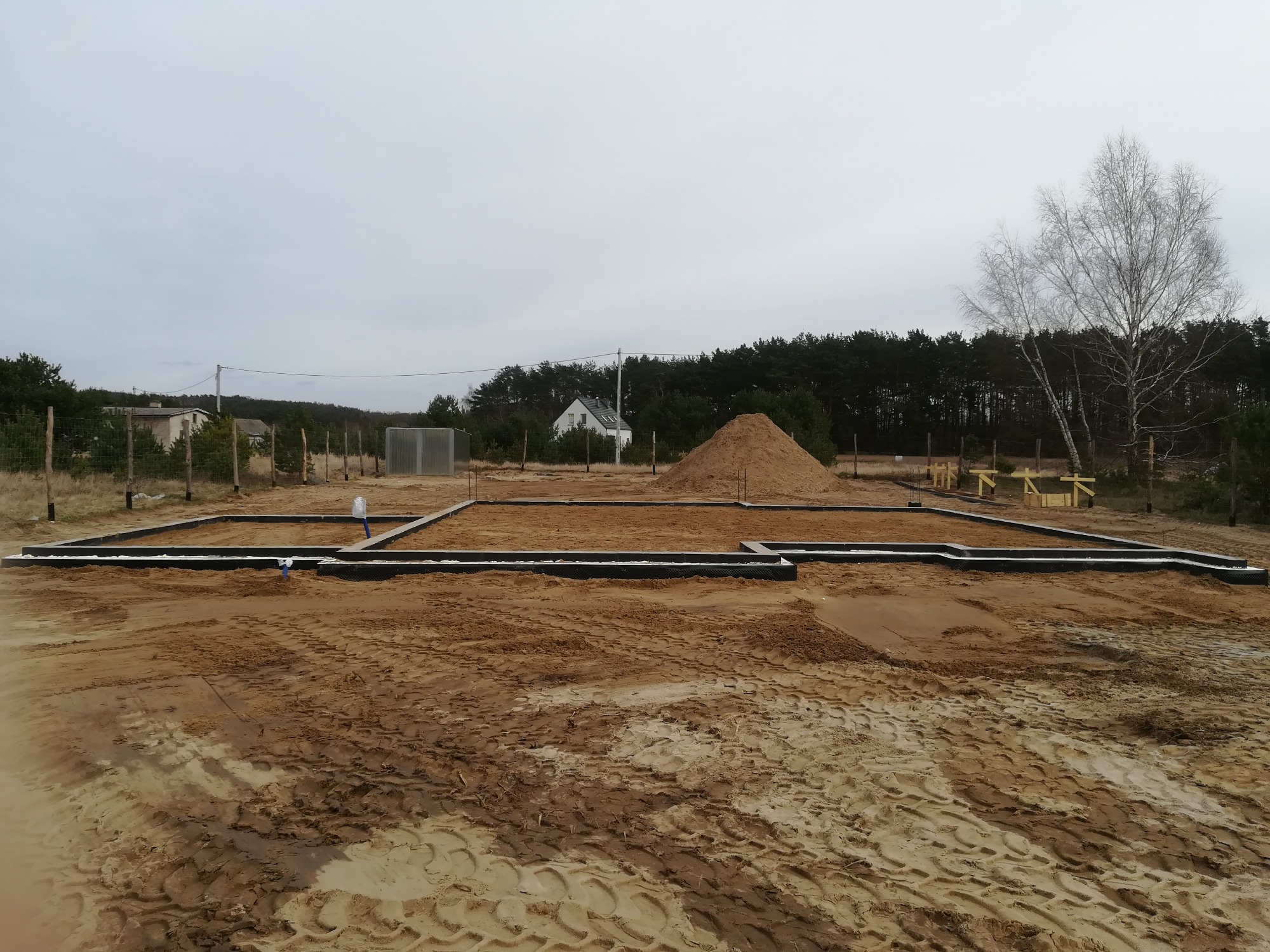 Przygotowany teren pod budowę domu, z wytyczonymi fundamentami i nasypaną górą piasku w tle, widok na las i pojedyncze budynki.