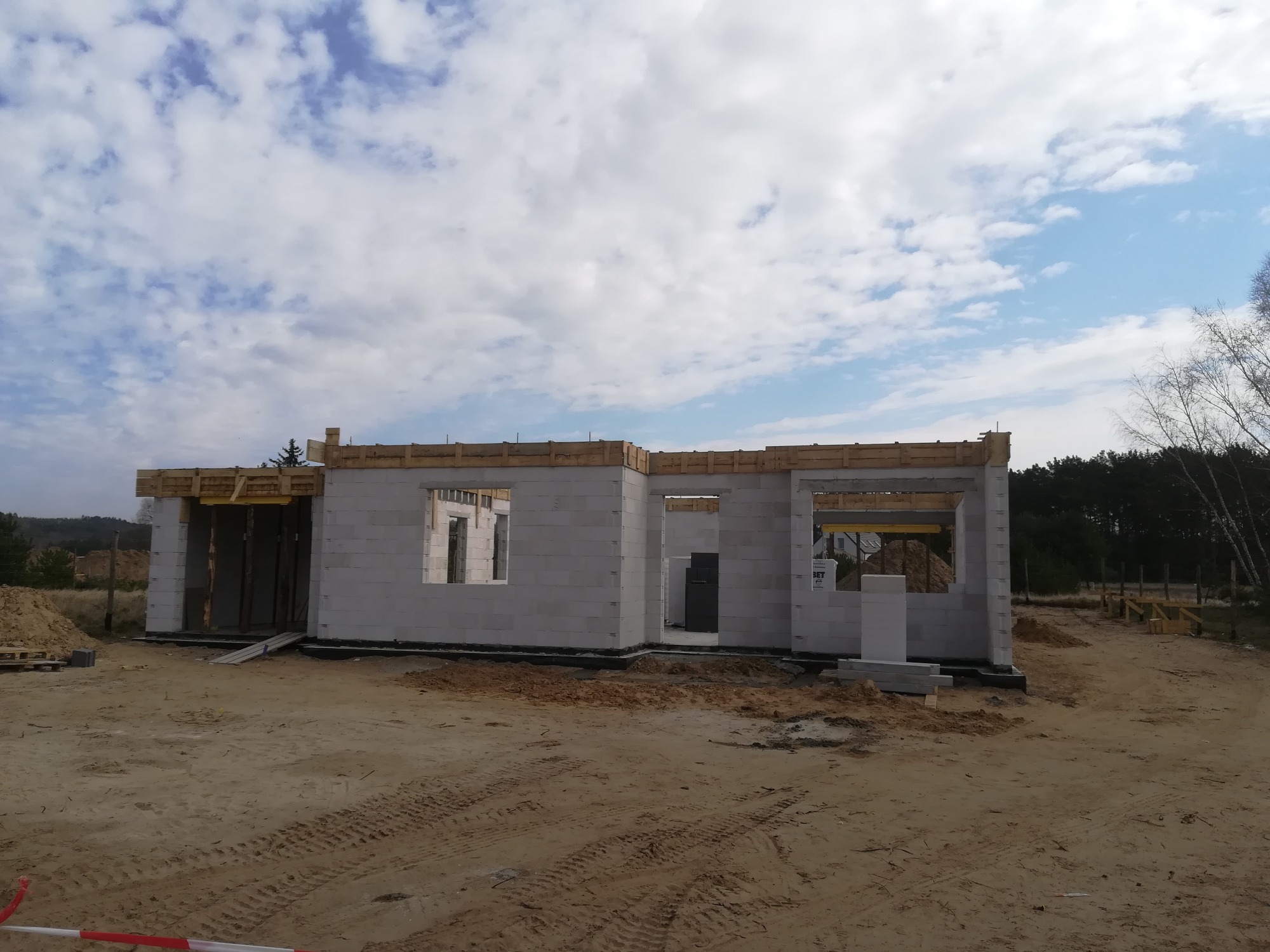 Wznoszenie ścian parterowego budynku z betonu komórkowego, widoczne drewniane szalunki nad otworami okiennymi i drzwiowymi, teren budowy z piaskiem i śladami opon.