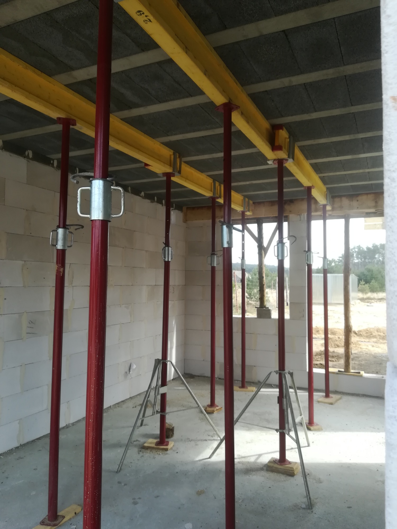 Wnętrze w trakcie budowy z widocznymi podporami stropowymi w kolorze czerwonym i żółtym, ustawionymi na drewnianych podkładkach, ściany z białych bloczków.