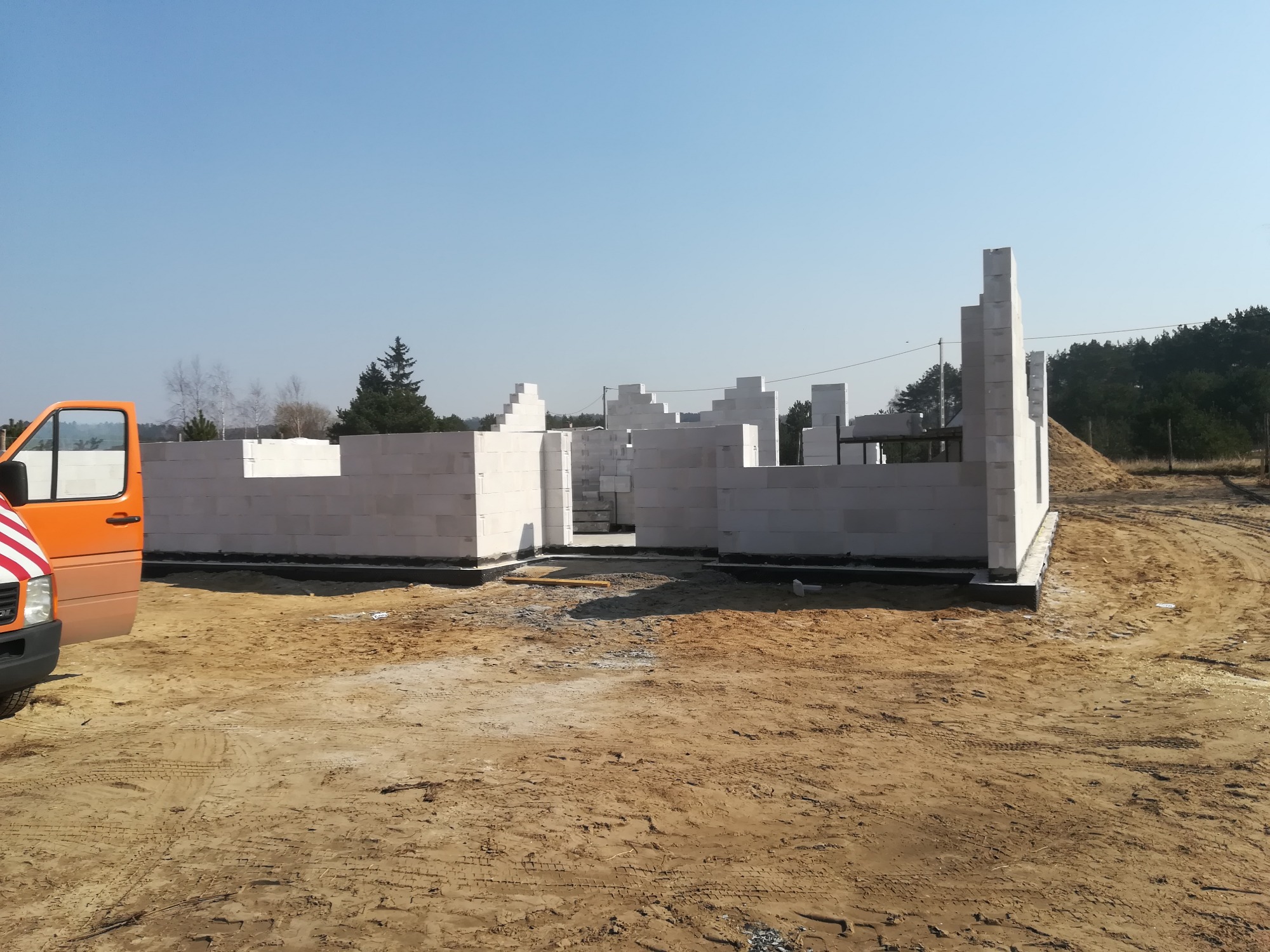 Wznoszone ściany parterowego budynku z białych bloczków na fundamencie, widoczny pomarańczowy samochód dostawczy, teren budowy z piaskiem i drzewami w tle.