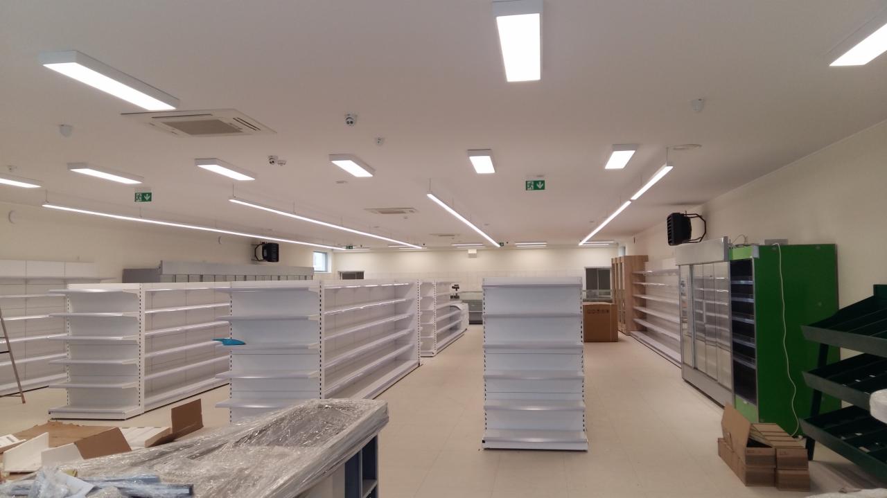 Wnętrze nowo wykończonego sklepu z zamontowanym oświetleniem LED, widoczne puste białe regały, system wentylacji pod sufitem oraz elementy chłodnicze.