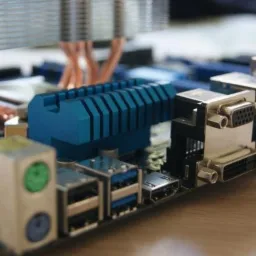 Zbliżenie na płytę główną komputera z widocznym układem chłodzenia z miedzianymi rurkami, niebieskim radiatorem i portami wejścia/wyjścia.