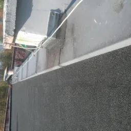 Płaski dach pokryty papą z widocznymi obróbkami blacharskimi, lina zabezpieczająca, drzewa w tle.