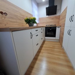 Primus - Biała kuchnia z drewnianymi akcentami: szafki z czarnymi uchwytami, drewniany blat i panel ścienny. Na blacie doniczka z żółtymi kwiatami. Nowoczesny design.