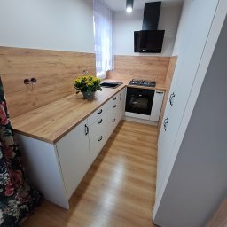 Primus - Nowoczesna kuchnia z białymi szafkami i drewnianym blatem. Na blacie stoi doniczka z żółtymi kwiatami. Widoczna płyta grzewcza i piekarnik.