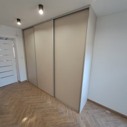 Primus - Beżowa szafa przesuwna na wymiar z trzema drzwiami, widoczna na tle parkietu ułożonego w jodełkę, oświetlona przez punktowe lampy sufitowe.