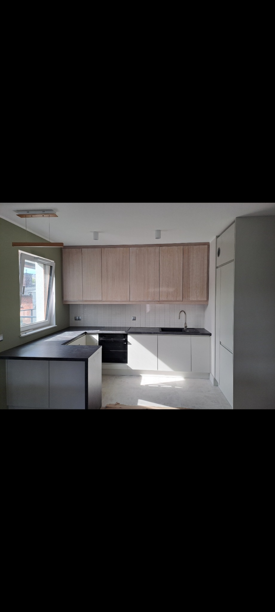 Jasne, nowoczesne meble kuchenne z drewnianymi górnymi szafkami i białymi dolnymi, czarny blat, widoczne okno z drewnianą lampą wiszącą, wpadające światło słoneczne.