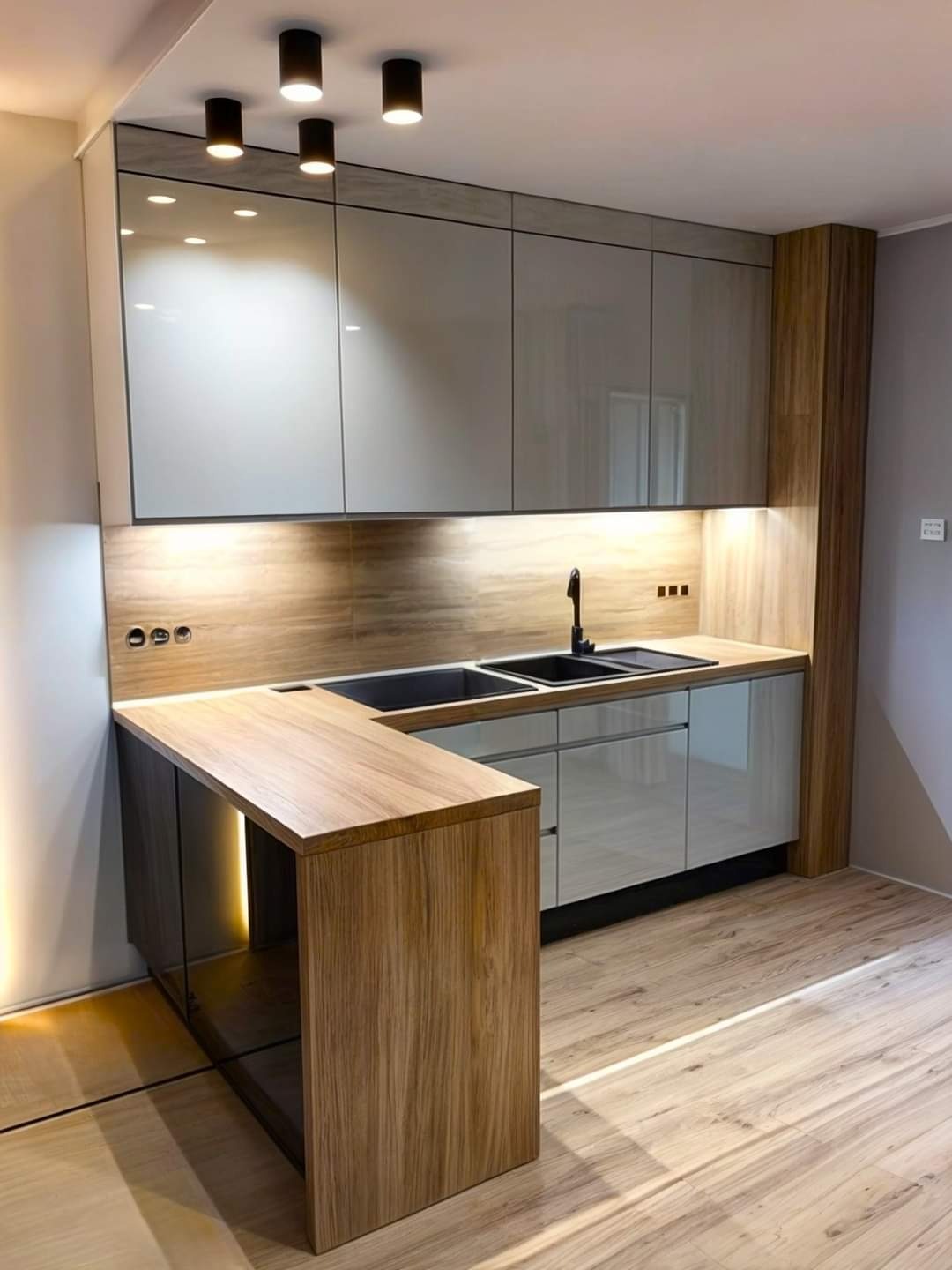 Nowoczesna kuchnia na wymiar z jasnymi, lakierowanymi frontami szafek, drewnianym blatem roboczym i czarną armaturą, oświetlona punktowymi lampami sufitowymi i listwą LED pod szafkami.