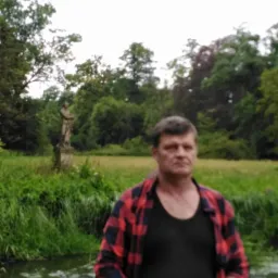 Mężczyzna w czerwonej koszuli w kratę na tle parku ze statuą i strumieniem. Portret w plenerze, naturalne światło, zieleń drzew i trawy.