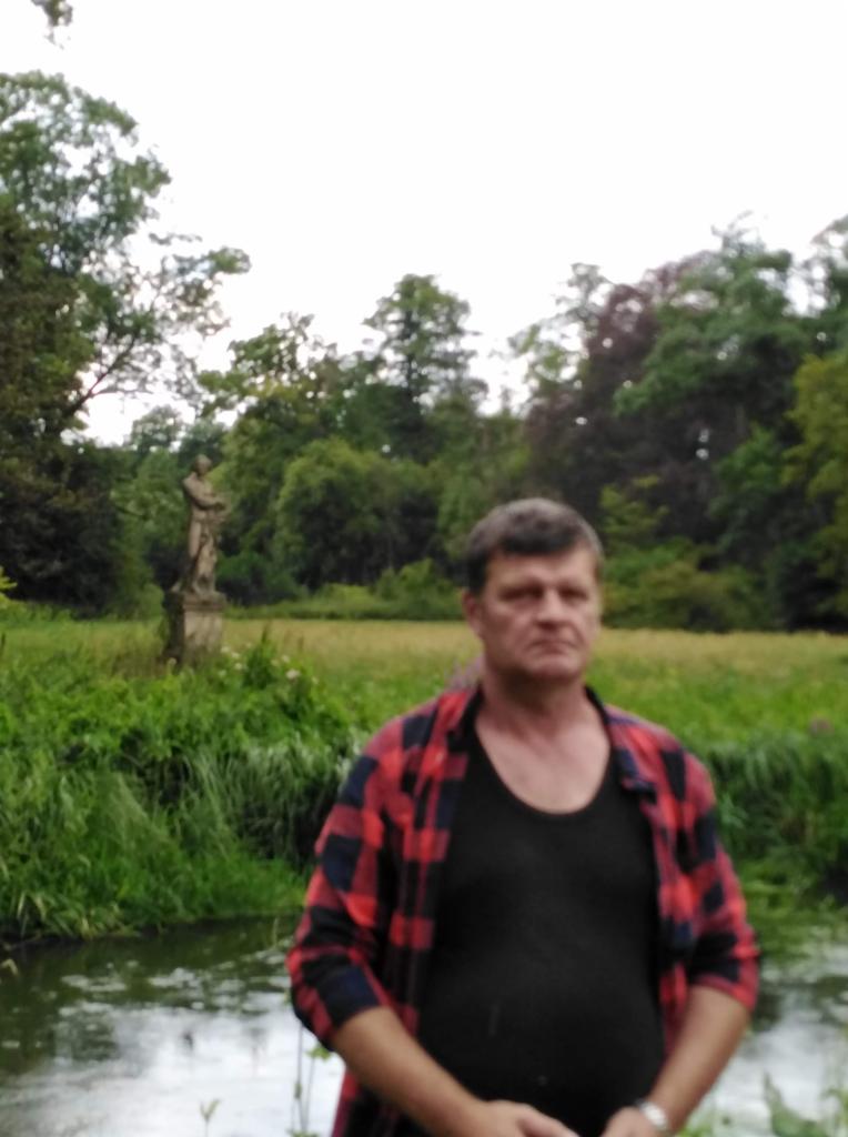 Mężczyzna w czerwonej koszuli w kratę na tle parku ze statuą i strumieniem. Portret w plenerze, naturalne światło, zieleń drzew i trawy.